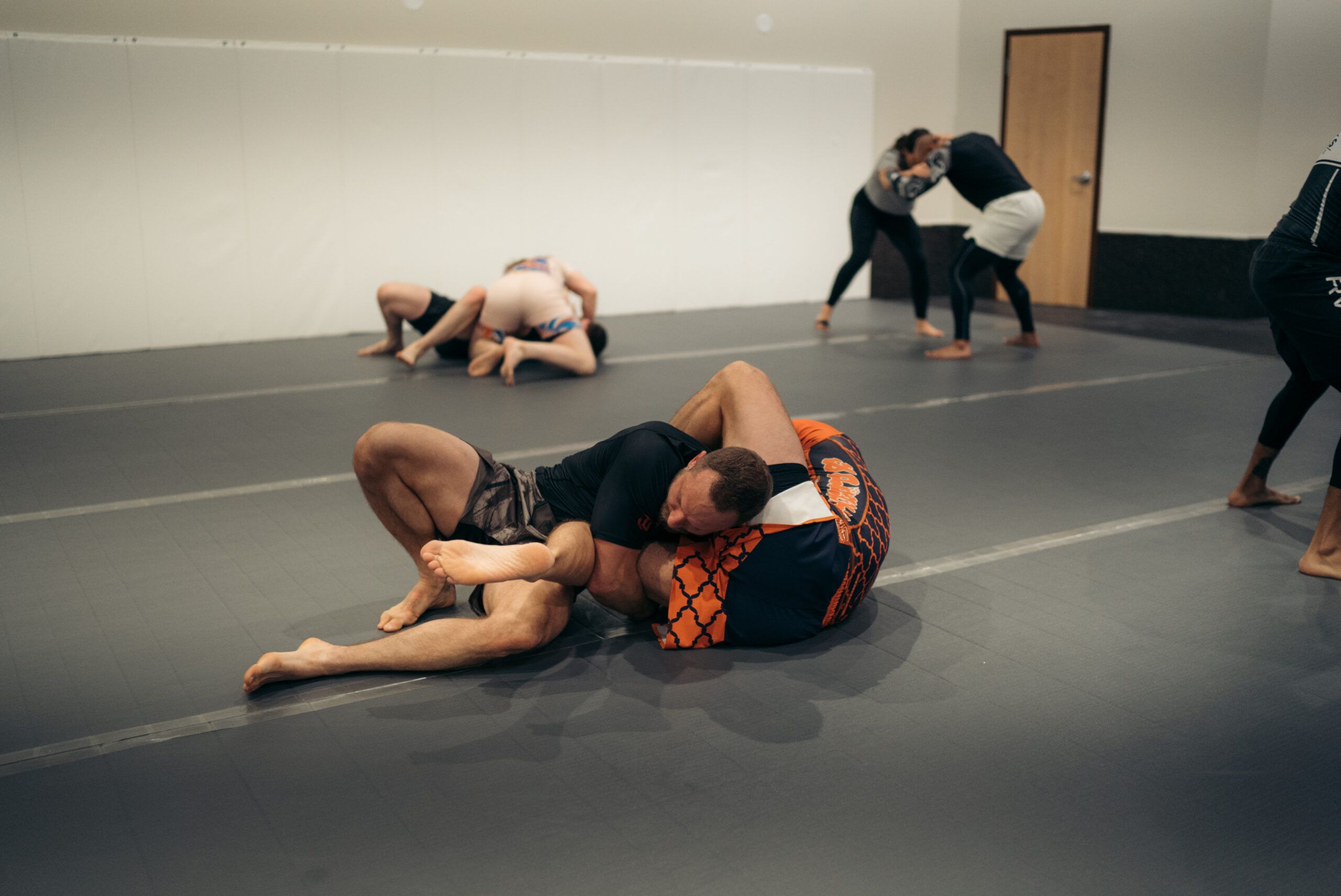 Two Atlas students rolling during a no-gi class while teammates train behind them